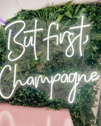 White neon sign reading 'But first, Champagne' mounted on a lush green foliage wall above a pink bench, trendy aesthetic photo backdrop
