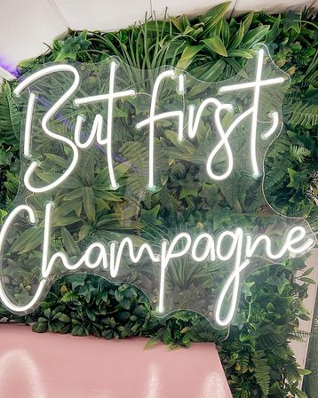White neon sign reading 'But first, Champagne' mounted on a lush green foliage wall above a pink bench, trendy aesthetic photo backdrop