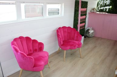 Two hot-pink scallop velvet chairs on light wood floor against a white shiplap wall, playful boutique lounge interior with a pink corrugated bar, disco ball, pink glass rack and neon 'But first Champagne' sign.