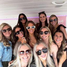 Smiling group of women in sunglasses taking a cheerful selfie under pink LED lights at a casual daytime gathering