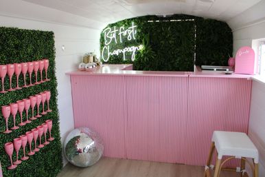 Pink pop-up champagne bar with ribbed counter, neon "But first Champagne" sign on a green plant wall, rows of pink champagne flutes displayed on a hedge, disco ball and wicker stool