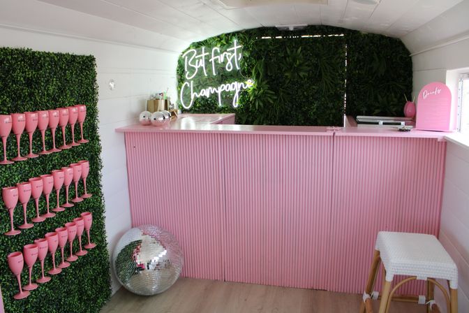 Pink pop-up champagne bar with ribbed counter, neon "But first Champagne" sign on a green plant wall, rows of pink champagne flutes displayed on a hedge, disco ball and wicker stool