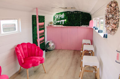Mobile pink champagne bar interior with neon 'But First Champagne' sign, scallop velvet chair, disco ball, faux greenery wall, ribbed pink counter and woven bar stools.