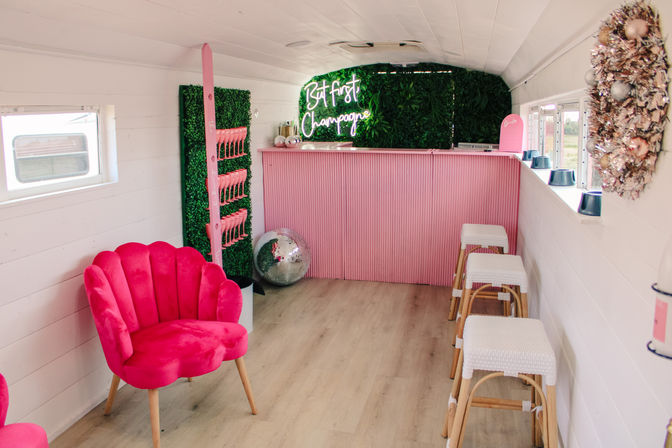 Mobile pink champagne bar interior with neon 'But First Champagne' sign, scallop velvet chair, disco ball, faux greenery wall, ribbed pink counter and woven bar stools.