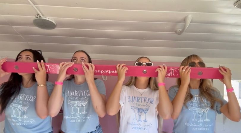 Four friends in matching light blue and white tees and pink wristbands leaning forward to drink from a long hot-pink shot-ski with cup holes inside a white-paneled room — playful group party scene.