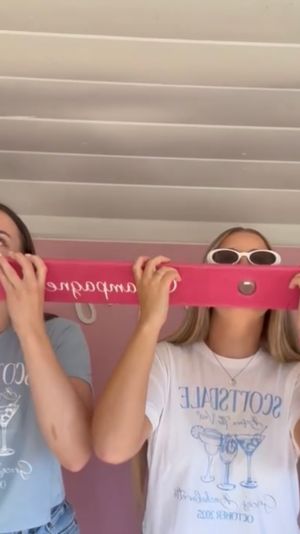 Four friends in matching light blue and white tees and pink wristbands leaning forward to drink from a long hot-pink shot-ski with cup holes inside a white-paneled room — playful group party scene.