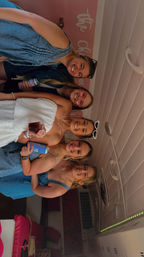 Five friends smiling in a group photo inside a pink-decorated indoor space, wearing summer dresses and sunglasses and holding drinks and can coolers — lively party vibe.