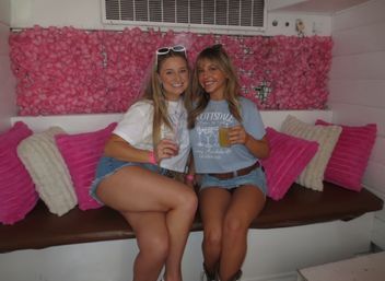 Two smiling young women in denim shorts holding drinks on a bench in front of a pink floral backdrop with bright pink pillows — casual indoor party photo-op.