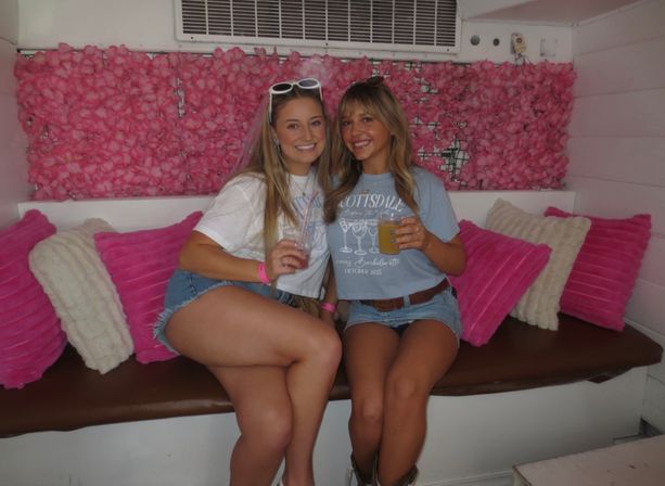 Two smiling young women in denim shorts holding drinks on a bench in front of a pink floral backdrop with bright pink pillows — casual indoor party photo-op.