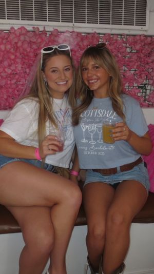 Two smiling young women in denim shorts holding drinks on a bench in front of a pink floral backdrop with bright pink pillows — casual indoor party photo-op.