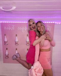 Two women in pink outfits smiling and hugging at an indoor pink-themed party photo booth with purple LED lights and champagne bottle backdrop