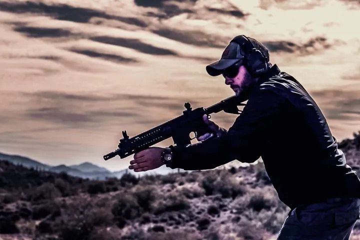 Person wearing a cap, sunglasses and hearing protection aiming a tactical rifle across scrubby desert hills at dusk.