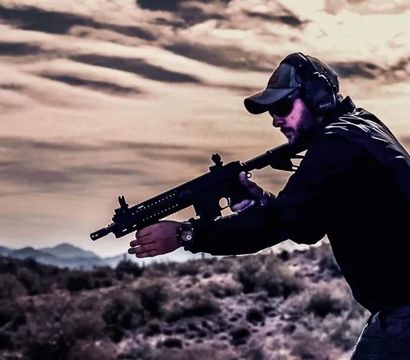 Person wearing a cap, sunglasses and hearing protection aiming a tactical rifle across scrubby desert hills at dusk.