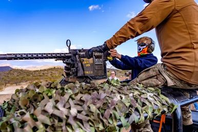 Outdoor desert shooting range: two people on an off‑road vehicle operating a mounted heavy machine gun draped in camouflage netting, one guiding while the other wears hearing protection and a colorful face mask under a bright blue sky.