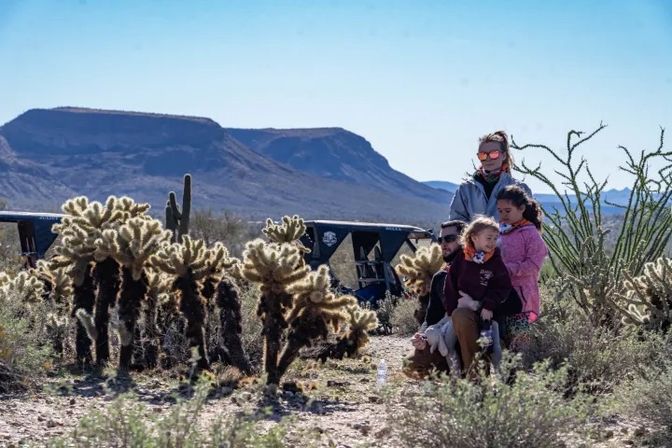 Group of adults and children sitting among spiky cholla cacti in a sunny Southwestern desert, off-road vehicle nearby and flat-topped mesas on the horizon