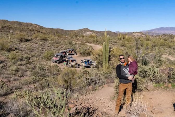 Adult holding a child on a rocky desert trail with parked off‑road UTVs, tall saguaro cacti and rolling desert hills under a clear blue sky — family outdoor adventure in the American Southwest.