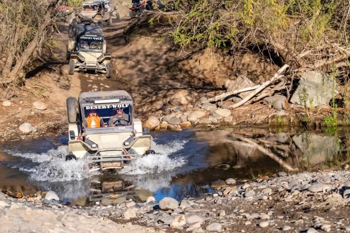 Off-road UTVs splashing through a shallow creek on a rocky dirt trail amid scrubby trees and sunlit riverbank