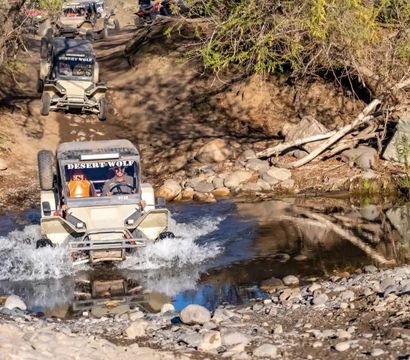 Off-road UTVs splashing through a shallow creek on a rocky dirt trail amid scrubby trees and sunlit riverbank