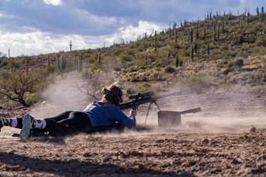 Shooter lying prone firing a scoped rifle on a dusty outdoor range in the Sonoran Desert, saguaro-studded hills and a cloudy sky in the background.