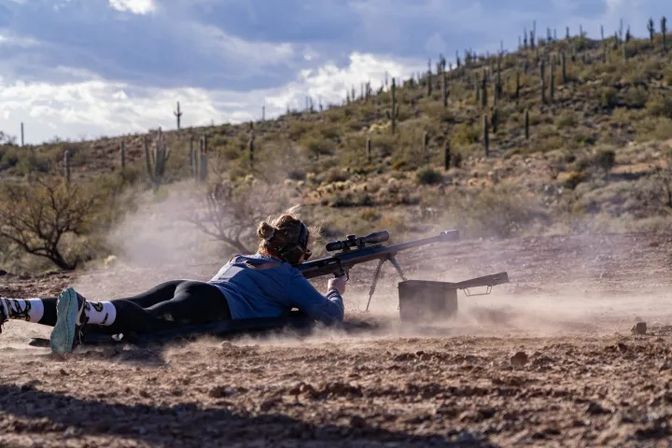 Shooter lying prone firing a scoped rifle on a dusty outdoor range in the Sonoran Desert, saguaro-studded hills and a cloudy sky in the background.