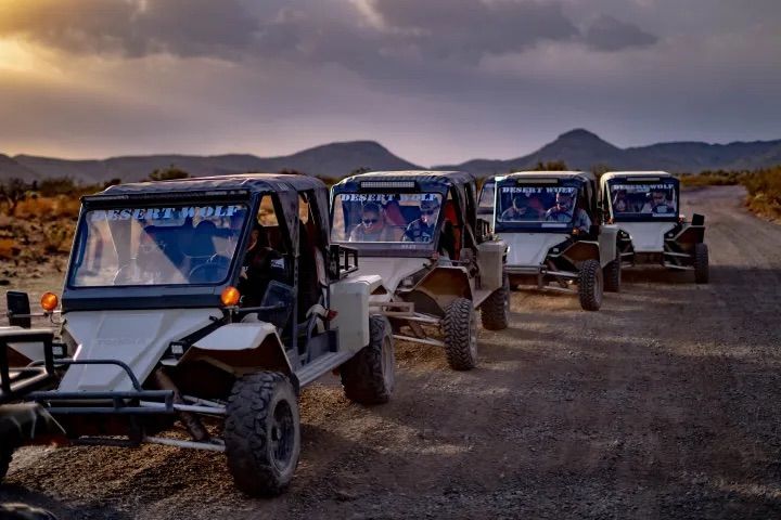 Four off-road UTV dune buggies in convoy on a dusty desert road at sunset, passengers visible inside, rugged mountains and dramatic clouds in the background — desert off-road adventure scene