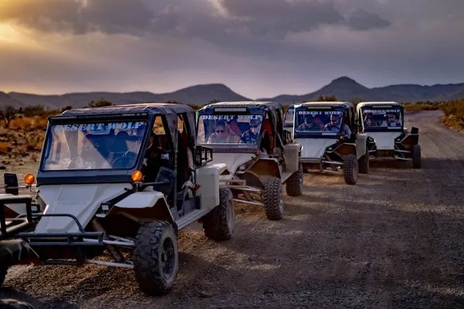 Line of four off-road UTV dune buggies on a dusty desert road at sunset with low mountains on the horizon and drivers visible through the windshields.