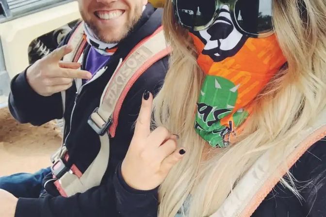Two people in an off-road UTV wearing safety harnesses — a grinning person flashing a peace sign and a long-blond-haired person with sunglasses and an orange-green bandana over their face making a rock-on gesture.