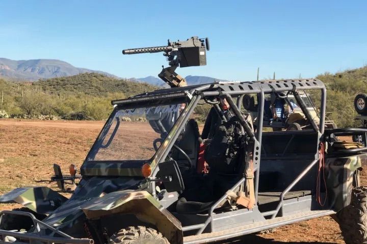 Camouflage off-road side-by-side UTV with rooftop-mounted heavy machine gun and roll cage parked on a desert trail with cacti and distant mountains under a clear blue sky.