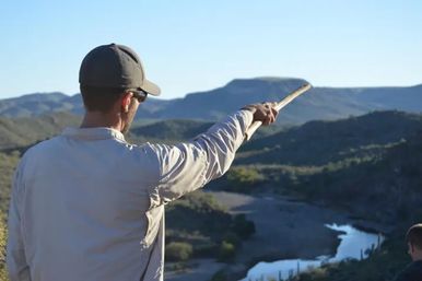 Hiker in a cap pointing with a stick toward a winding river and layered mountain ridges in a sunlit desert valley