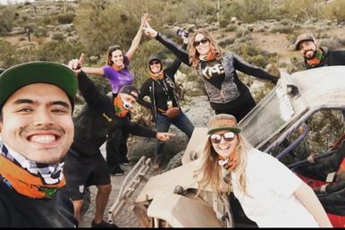 Seven people posing for a selfie around a dusty off-road buggy on a rocky desert trail, wearing caps, sunglasses and colorful neck gaiters with scrubby hills in the background.