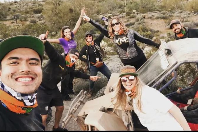 Seven people posing for a selfie around a dusty off-road buggy on a rocky desert trail, wearing caps, sunglasses and colorful neck gaiters with scrubby hills in the background.