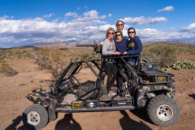 Four adults posing on a camouflage off-road UTV side-by-side in a desert landscape with cacti and distant mountains under a bright blue sky with scattered clouds.