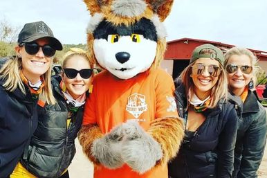 Four smiling women in sunglasses and bandanas pose outdoors with a friendly wolf mascot in an orange shirt, standing in front of a red barn in a desert setting.