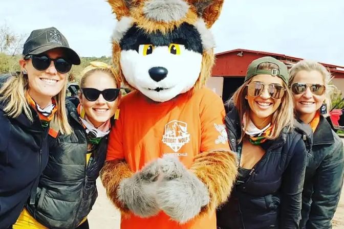 Four smiling women in sunglasses and bandanas pose outdoors with a friendly wolf mascot in an orange shirt, standing in front of a red barn in a desert setting.
