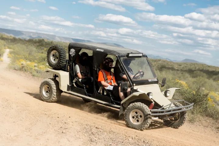 Off-road dune buggy kicking up dust on a desert trail with three helmeted riders, scrub vegetation and distant mountains under a blue sky.