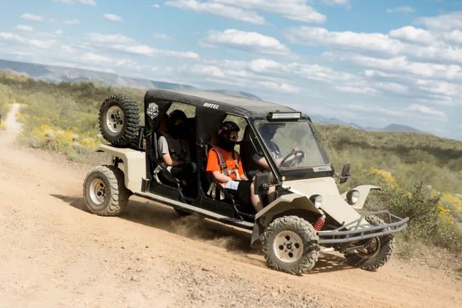 Off-road dune buggy kicking up dust on a desert trail with three helmeted riders, scrub vegetation and distant mountains under a blue sky.