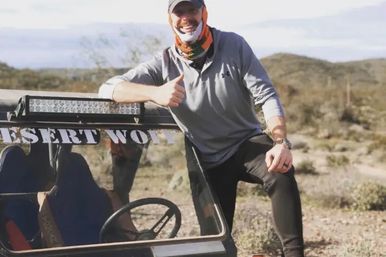Smiling person in a cap and neck gaiter giving a thumbs-up while leaning on an off-road buggy in a sunlit desert landscape, evoking outdoor adventure and rugged terrain.