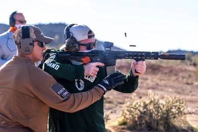 Two people at an outdoor desert shooting range: an instructor guiding a shooter firing a modern rifle with ear and eye protection, a spent shell casing ejecting midair.