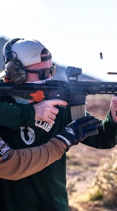 Two people at an outdoor desert shooting range: an instructor guiding a shooter firing a modern rifle with ear and eye protection, a spent shell casing ejecting midair.