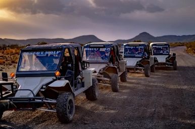 Four off-road dune buggies lined up on a dusty desert trail at sunset, passengers inside, rugged mountains and a dramatic sky in the background.
