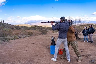 Person firing a rifle with ear protection at an outdoor desert shooting range, instructor steadying the stance as shell casings fly, cacti and scrub with distant mountains under a bright blue sky and a few spectators watching.