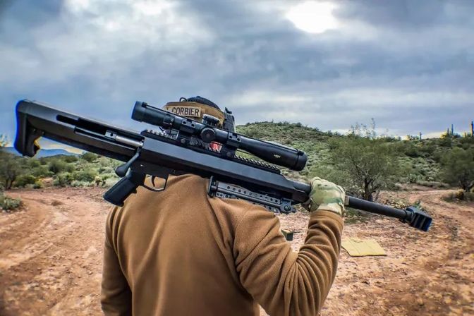 Person carrying a long-range precision rifle with scope on their shoulder, wearing a tan jacket and camo gloves, walking along a cactus-studded desert trail under a cloudy sky.