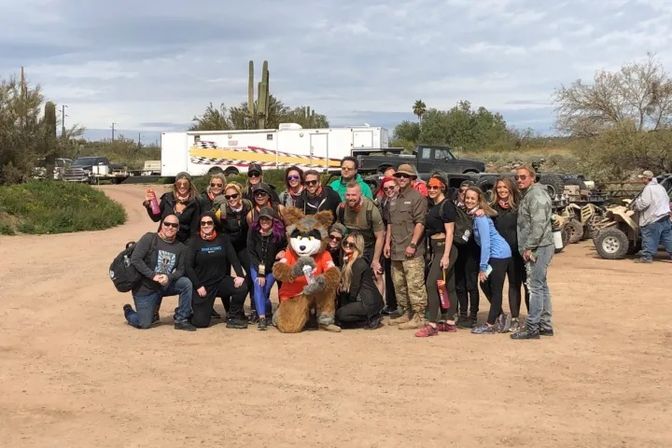 Group of about 20 adults posing with a costumed fox mascot in a Southwestern desert staging area with ATVs, trailers and saguaro cacti under a cloudy sky