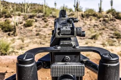 Close-up view down the iron sights of a mounted machine gun on a tripod, aiming over a sunlit arid desert dotted with tall cacti.