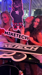 Three people smiling under pink and purple party lights, posing with a large cartoon DJ bear cutout and a heart-shaped promotional sign at an indoor nightlife event.