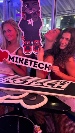 Three people smiling under pink and purple party lights, posing with a large cartoon DJ bear cutout and a heart-shaped promotional sign at an indoor nightlife event.
