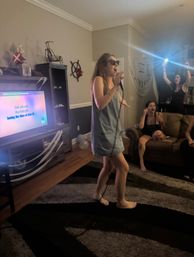 Barefoot woman in a casual dress and sunglasses singing into a microphone at a living-room karaoke party, friends cheering and recording with a phone while lyrics play on the TV.