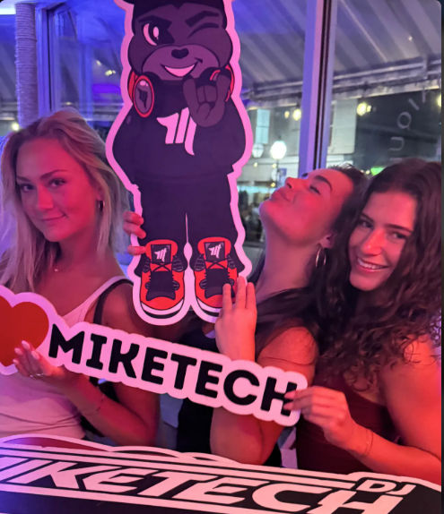 Three friends posing with a cartoon bear mascot cutout and heart-shaped sign in a pink-purple neon-lit bar or nightclub, smiling and playful.