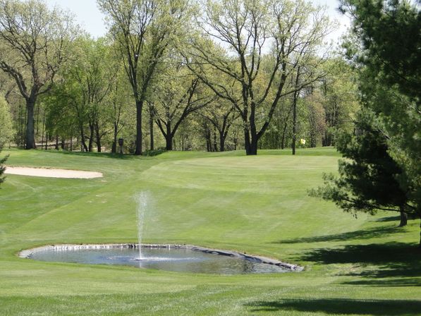 Lush golf course green with a small pond and fountain in the foreground, sand bunker to the left and a flag on the distant green framed by mature trees.