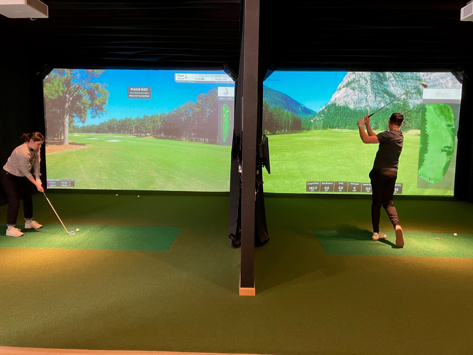 Two golfers in side-by-side indoor golf simulator bays practicing swings on green turf mats with large projected fairway screens showing a virtual course and shot distance stats.